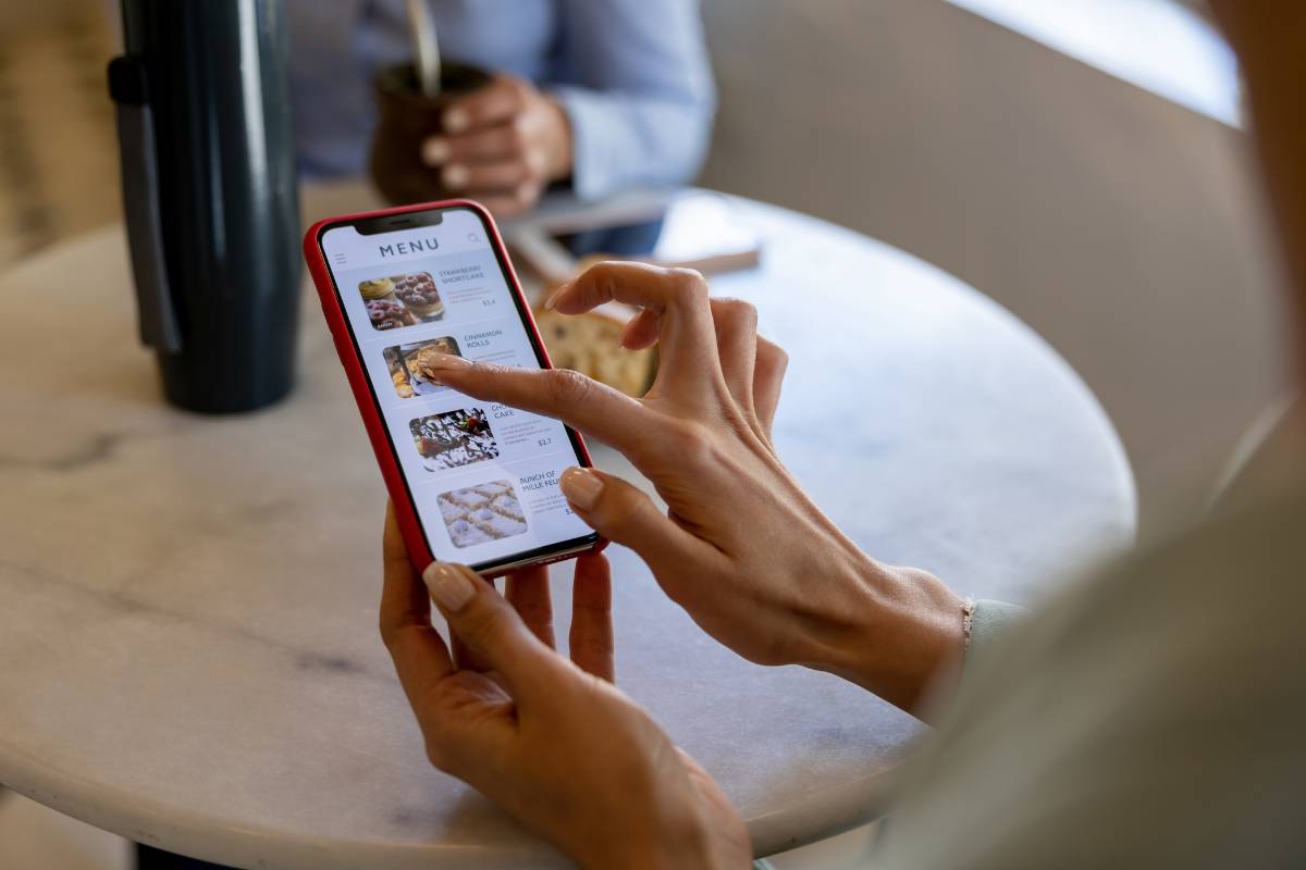 Mão de uma pessoa navegando no cardápio digital de um restaurante em seu celular, exibindo imagens e descrições dos pratos.