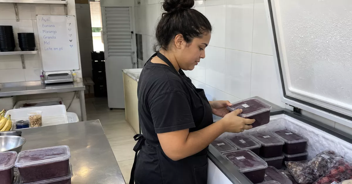 Funcionária organiza recipientes de açaí dentro de um freezer horizontal aberto na área limpa de armazenamento da açaiteria.