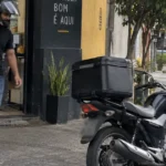 Entregador sai de uma hamburgueria com o pedido e caminha em direção à motocicleta.