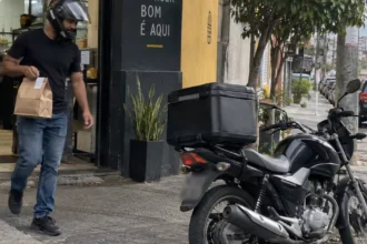 Entregador sai de uma hamburgueria com o pedido e caminha em direção à motocicleta.