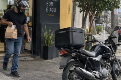Entregador sai de uma hamburgueria com o pedido e caminha em direção à motocicleta.