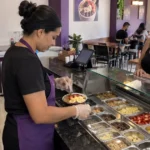 Funcionária monta uma tigela de açaí com frutas e complementos diante de uma cliente no balcão de uma açaiteria.