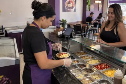 Funcionária monta uma tigela de açaí com frutas e complementos diante de uma cliente no balcão de uma açaiteria.