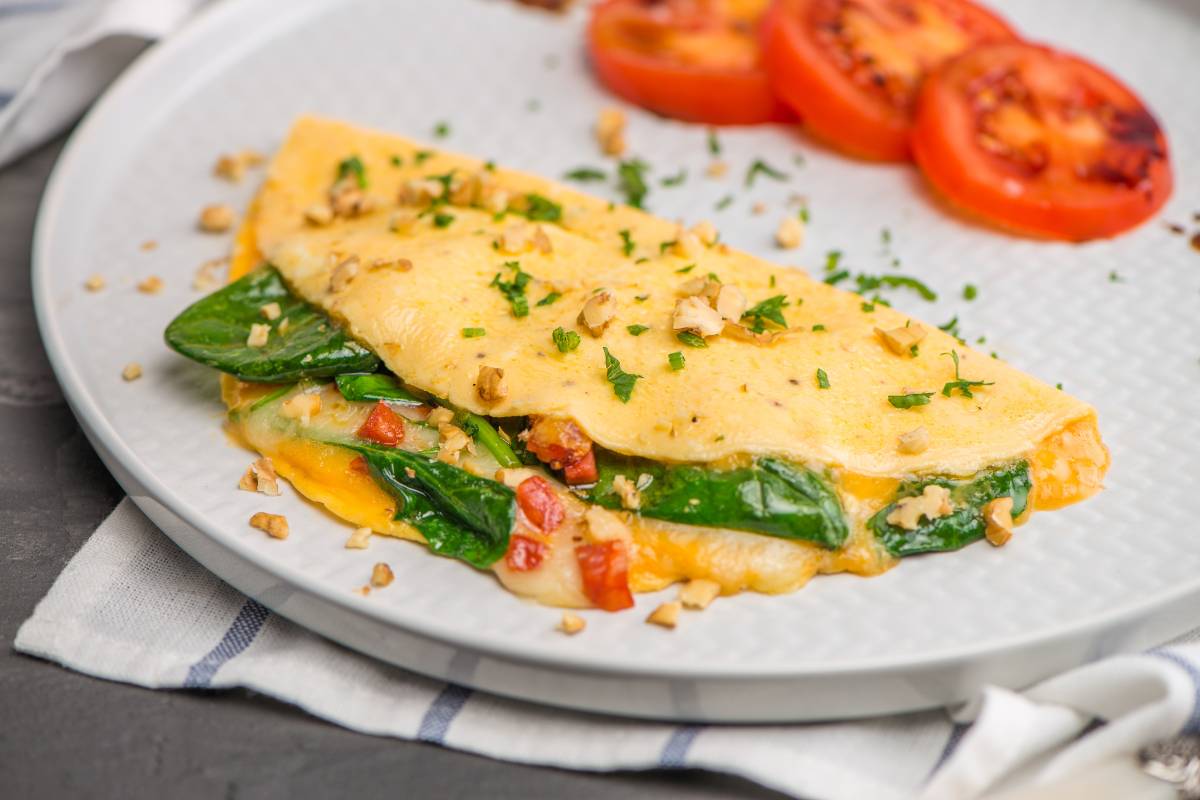 Omelete low carb recheado com espinafre e queijo: Omelete dourado recheado com espinafre, queijo e tomate, decorado com ervas frescas e nozes picadas, acompanhado de tomate grelhado.