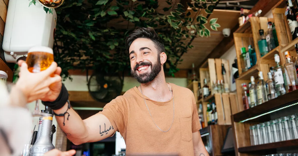 Proprietário de bar entrega um copo de chope ao cliente diante do balcão, com bebidas e prateleiras ao fundo.