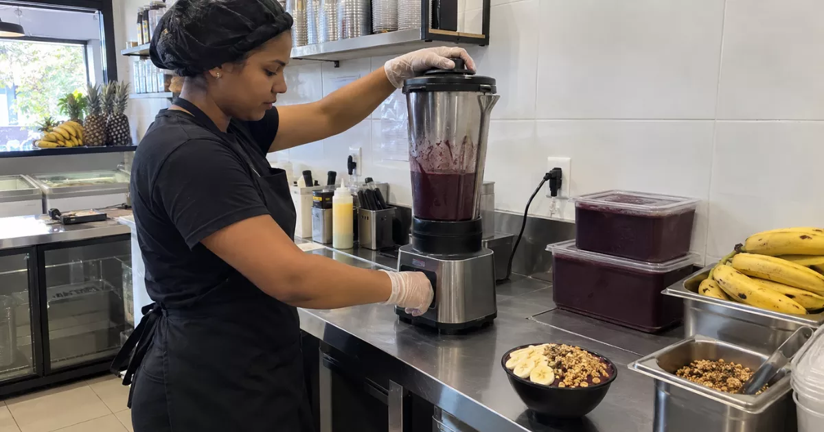 Funcionária prepara açaí em liquidificador industrial sobre bancada de inox, com bananas, recipientes de polpa e tigela pronta ao lado.