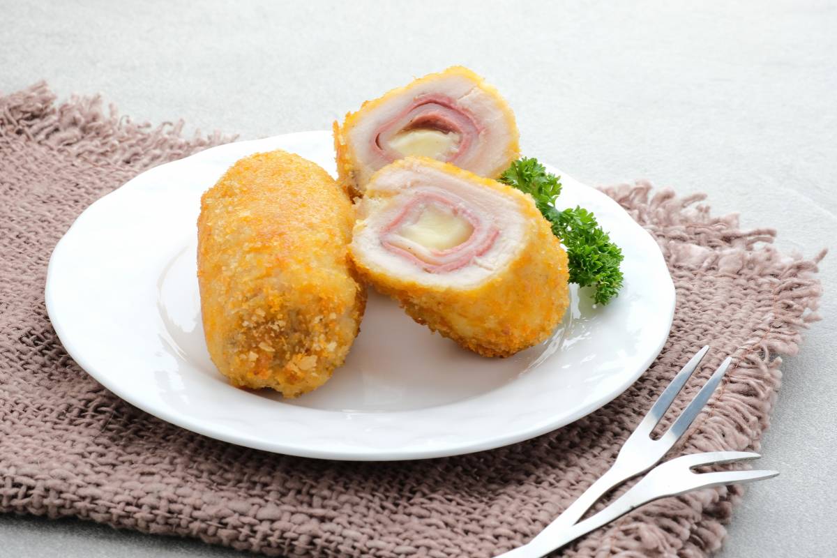 Enroladinho de frango com queijo e presunto: Enroladinho de frango recheado com presunto e queijo, servido empanado e cortado ao meio, com salsinha ao lado.