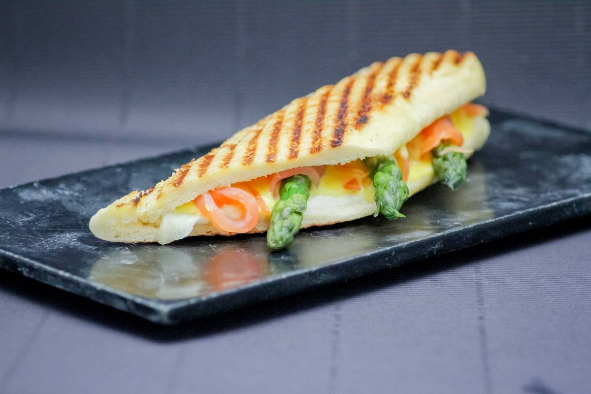 Panini low carb com salmão e aspargos: Sanduíche grelhado de panini low carb recheado com salmão defumado, aspargos e queijo, servido sobre uma placa de ardósia.