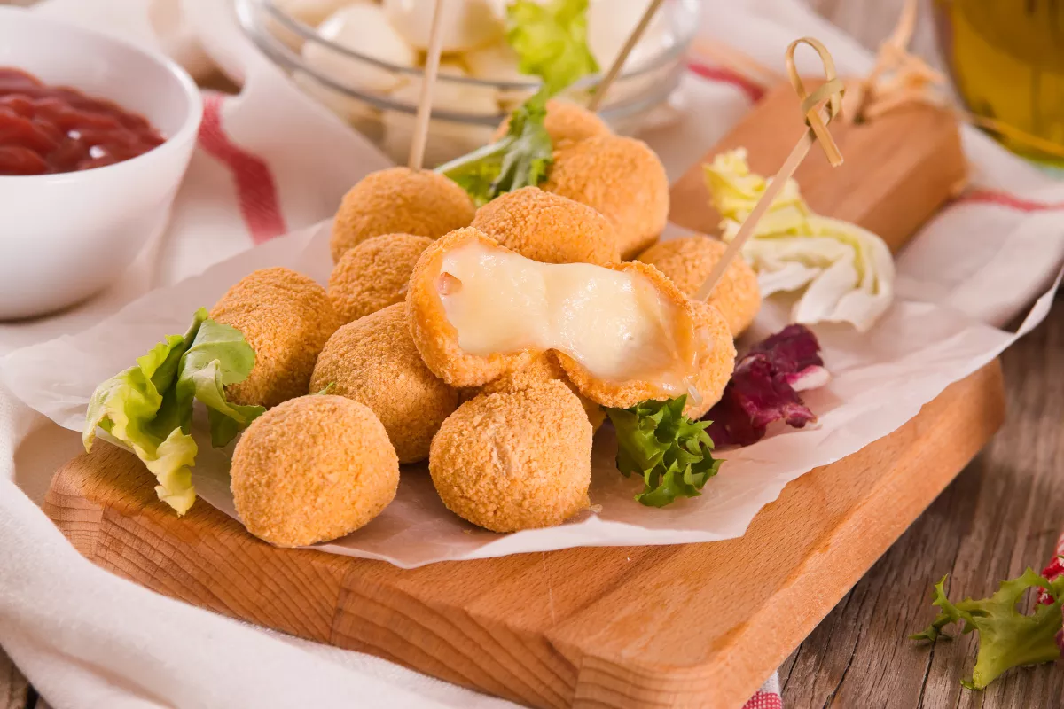Bolinho de queijo com recheio cremoso: Porção de bolinhos crocantes recheados com queijo derretido, servidos em uma tábua com folhas verdes e molhos para acompanhamento.