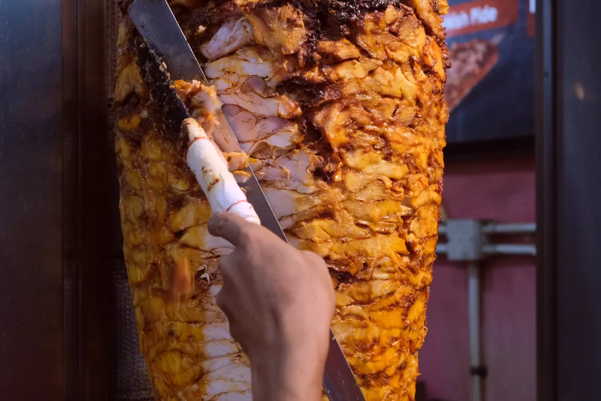 Mão cortando carne em uma churrasqueira vertical para churrasco grego, destacando o preparo suculento e a técnica de corte.