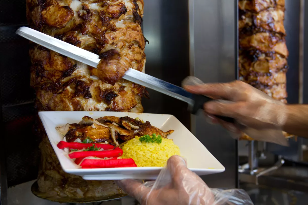Carne de churrasco grego sendo servida em um prato com arroz amarelo, picles e ervas frescas, mostrando uma apresentação atrativa.
