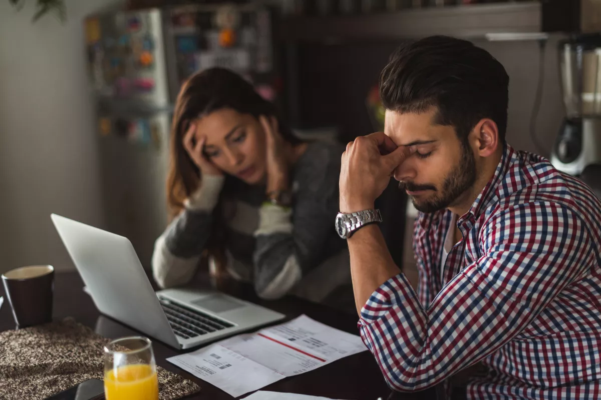 Casal sentado à mesa de casa, com expressões tensas enquanto olham para boletos e um laptop. Representa a dificuldade de clientes que enfrentam problemas financeiros para quitar suas dívidas.
