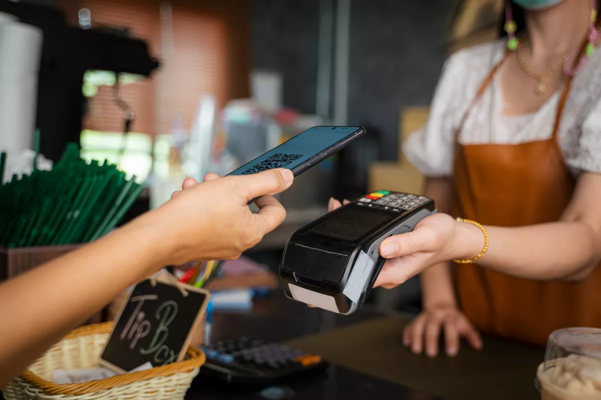Cliente realizando pagamento digital via QR Code em um estabelecimento. A atendente segura uma máquina de cartão enquanto o cliente aproxima o celular para concluir a transação.