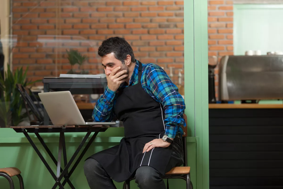 Homem vestindo avental sentado em uma cafeteria, olhando para o notebook com expressão de preocupação. Representa um empresário enfrentando dificuldades financeiras ou inadimplência de clientes.