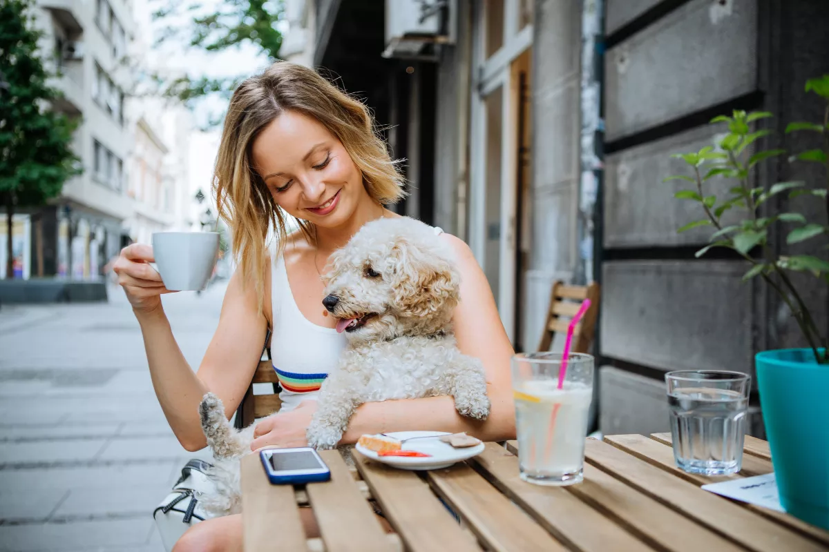 Uma mulher loira, sorridente, segura seu cachorro no colo enquanto toma café em um restaurante ao ar livre. A mesa tem um prato com um lanche e um copo de água com canudo. O cenário urbano ao fundo sugere um ambiente casual e pet friendly, ideal para clientes que querem desfrutar de momentos com seus animais de estimação.