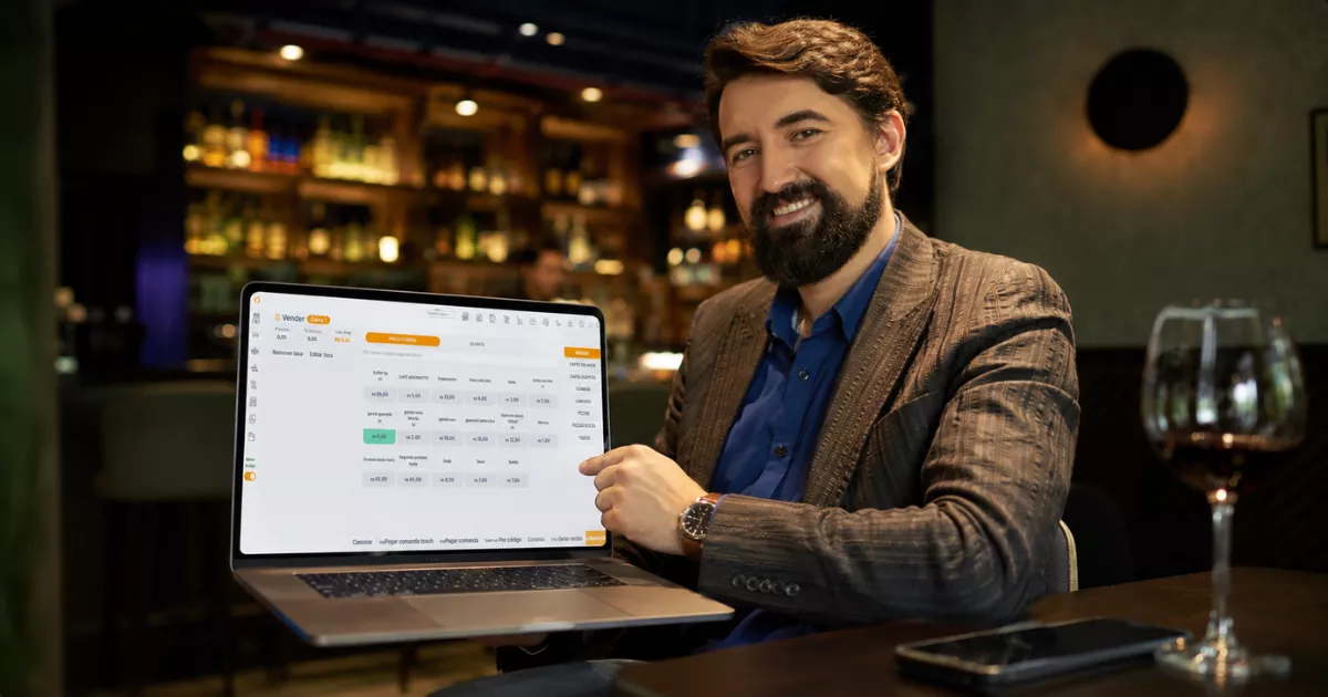 Dono de bar sorridente apresenta notebook com sistema de vendas ControleNaMão na tela, sentado em ambiente noturno com balcão iluminado ao fundo