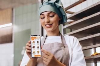 Confeiteira mostra no celular o PDV do ControleNaMão com produtos de doceria diante das assadeiras da área de produção.