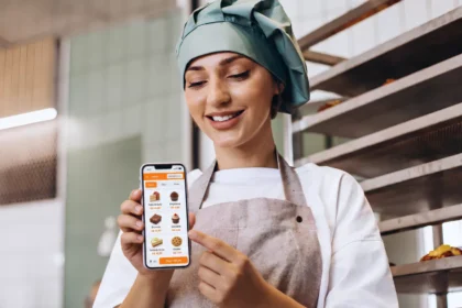 Confeiteira mostra no celular o PDV do ControleNaMão com produtos de doceria diante das assadeiras da área de produção.