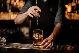 Bartender preparando um coquetel em copo de vidro com gelo, usando uma colher bailarina, atrás de um balcão profissional iluminado em bar noturno sofisticado.