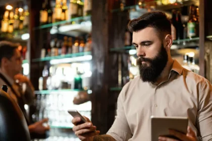 Gestor de bar consulta celular e tablet atrás do balcão, cercado por garrafas e prateleiras, durante a operação de um bar noturno.