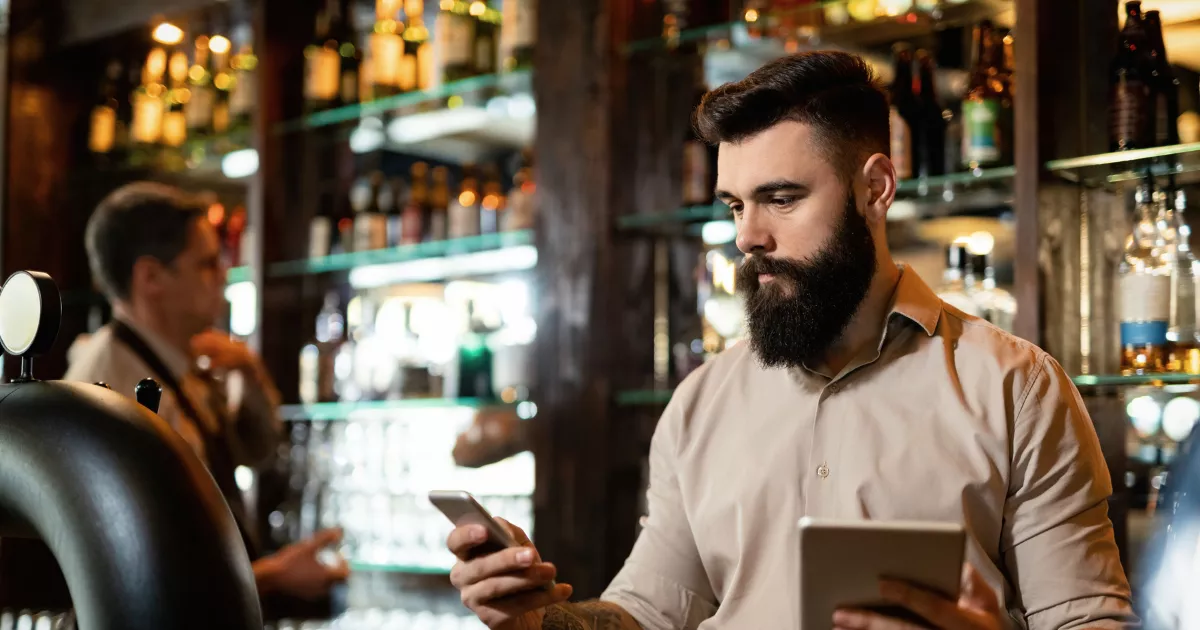 Gestor de bar consulta celular e tablet atrás do balcão, cercado por garrafas e prateleiras, durante a operação de um bar noturno.