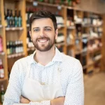 Homem sorridente usando avental branco dentro de uma distribuidora de bebidas bem organizada, com prateleiras repletas de garrafas variadas ao fundo. A imagem transmite profissionalismo e simpatia, ilustrando o perfil de um empreendedor que está começando seu negócio no setor de bebidas.