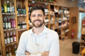 Homem sorridente usando avental branco dentro de uma distribuidora de bebidas bem organizada, com prateleiras repletas de garrafas variadas ao fundo. A imagem transmite profissionalismo e simpatia, ilustrando o perfil de um empreendedor que está começando seu negócio no setor de bebidas.
