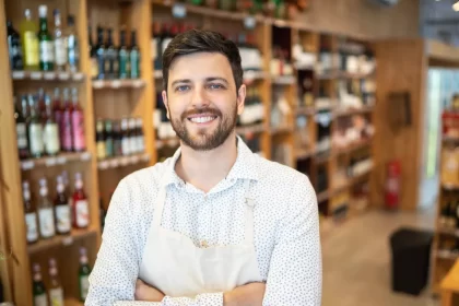 Homem sorridente usando avental branco dentro de uma distribuidora de bebidas bem organizada, com prateleiras repletas de garrafas variadas ao fundo. A imagem transmite profissionalismo e simpatia, ilustrando o perfil de um empreendedor que está começando seu negócio no setor de bebidas.