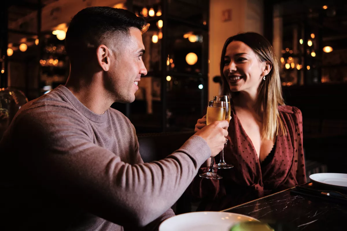 Casal sorridente brindando com taças de espumante em ambiente romântico e iluminado, sentado à mesa de um restaurante elegante. Representa o clima intimista e as experiências gastronômicas oferecidas no Dia dos Namorados.