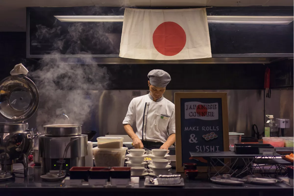 Chef japonês preparando sushi em estação culinária com bandeira do Japão ao fundo e utensílios de cozinha à frente. Homenagem à influência da imigração japonesa na gastronomia brasileira, especialmente em restaurantes orientais.

