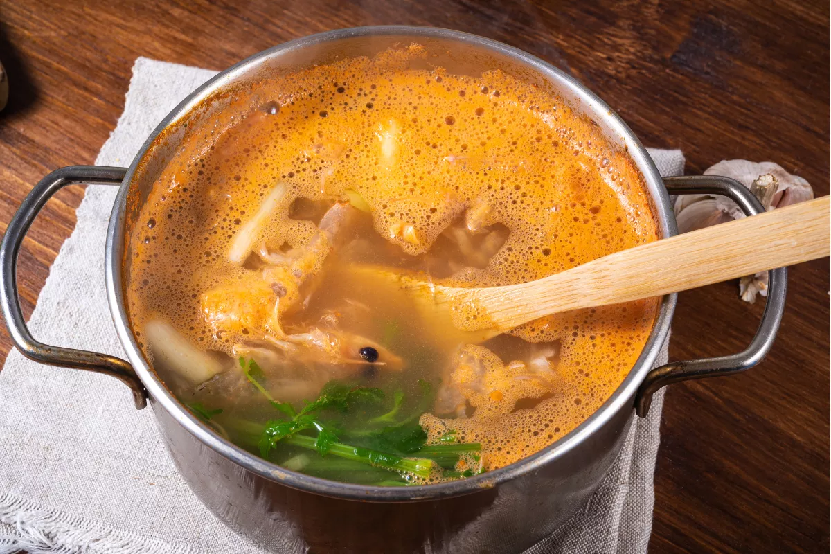 Panela de sopa fumegante com caldo grosso e pedaços de carne, sendo mexida com colher de pau sobre um pano rústico. Representa o início do inverno e o aumento da procura por pratos quentes e aconchegantes em restaurantes.