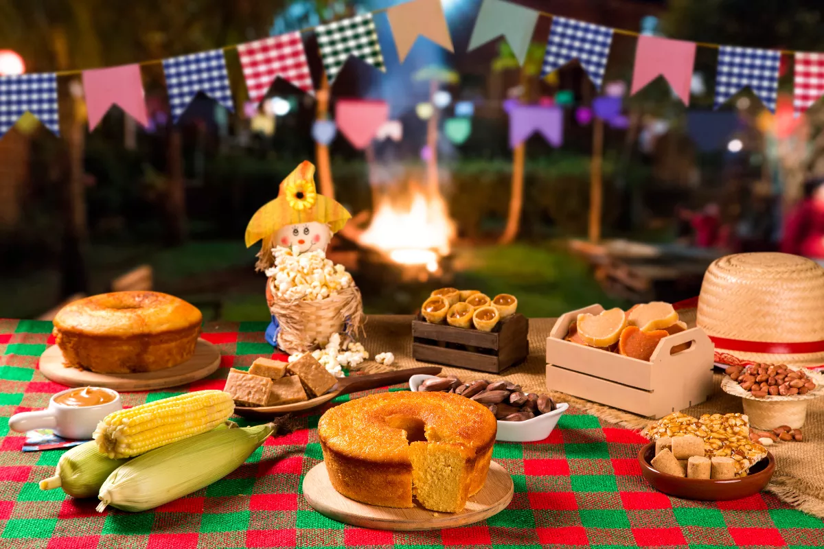 Mesa decorada com toalha xadrez e comidas típicas juninas, como bolo de milho, pamonha, pipoca, pé de moleque e milho cozido, em frente a uma fogueira e bandeirinhas coloridas. Celebração tradicional de São João e destaque para menus temáticos em junho.
