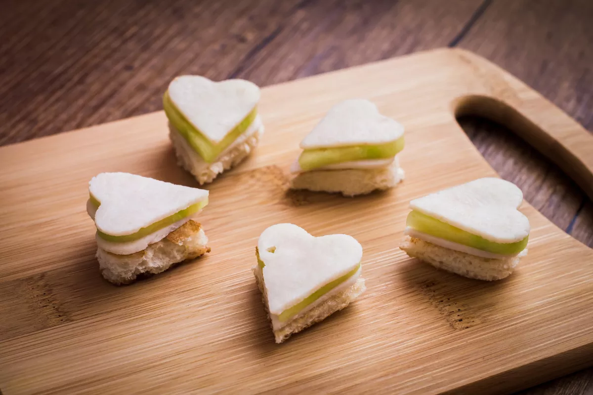 Mini sanduíches em formato de coração com camadas de pão e pepino, sobre tábua de madeira, perfeitos para entrada temática com apelo visual no Dia das Mães.