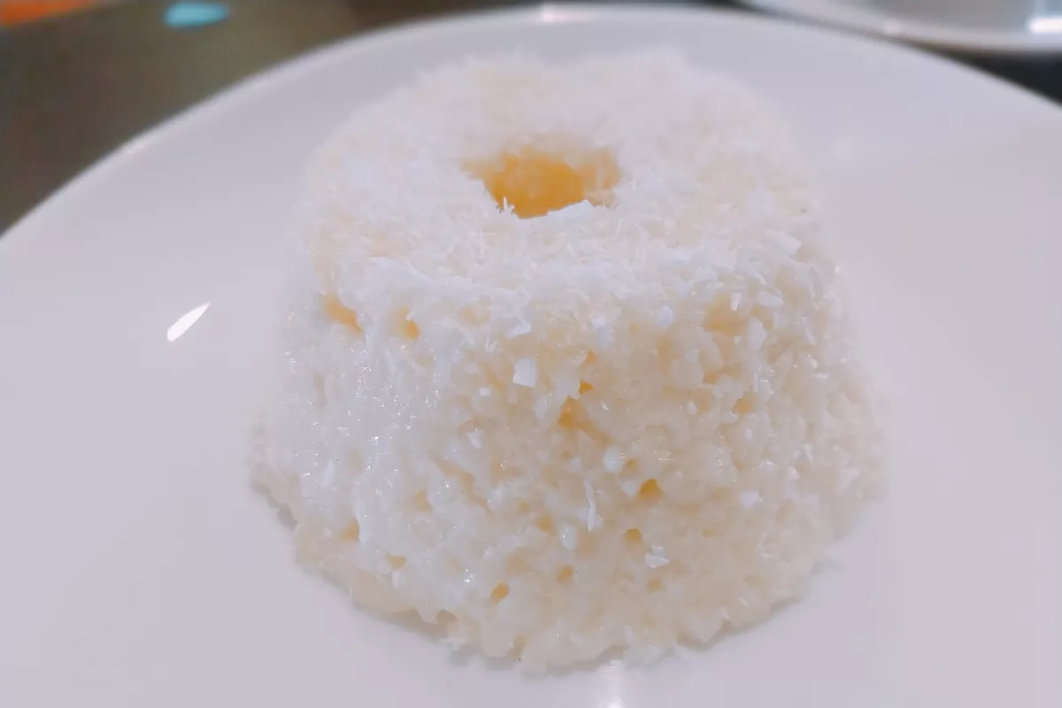 Bolo individual de tapioca com coco servido em prato branco, com textura úmida e acabamento com coco ralado, evocando memórias afetivas e sabores regionais.