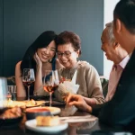 Família asiática reunida em restaurante, brindando com taças de vinho e sorrindo ao redor da mesa, representando o clima afetivo e acolhedor da data comemorativa.