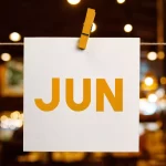 Cartão branco pendurado com prendedor de madeira exibindo a palavra “JUN” em letras grandes e alaranjadas, com luzes desfocadas ao fundo. Representa o mês de junho como um todo, com foco em planejamento de ações sazonais para bares e restaurantes.