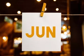 Cartão branco pendurado com prendedor de madeira exibindo a palavra “JUN” em letras grandes e alaranjadas, com luzes desfocadas ao fundo. Representa o mês de junho como um todo, com foco em planejamento de ações sazonais para bares e restaurantes.