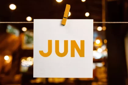 Cartão branco pendurado com prendedor de madeira exibindo a palavra “JUN” em letras grandes e alaranjadas, com luzes desfocadas ao fundo. Representa o mês de junho como um todo, com foco em planejamento de ações sazonais para bares e restaurantes.