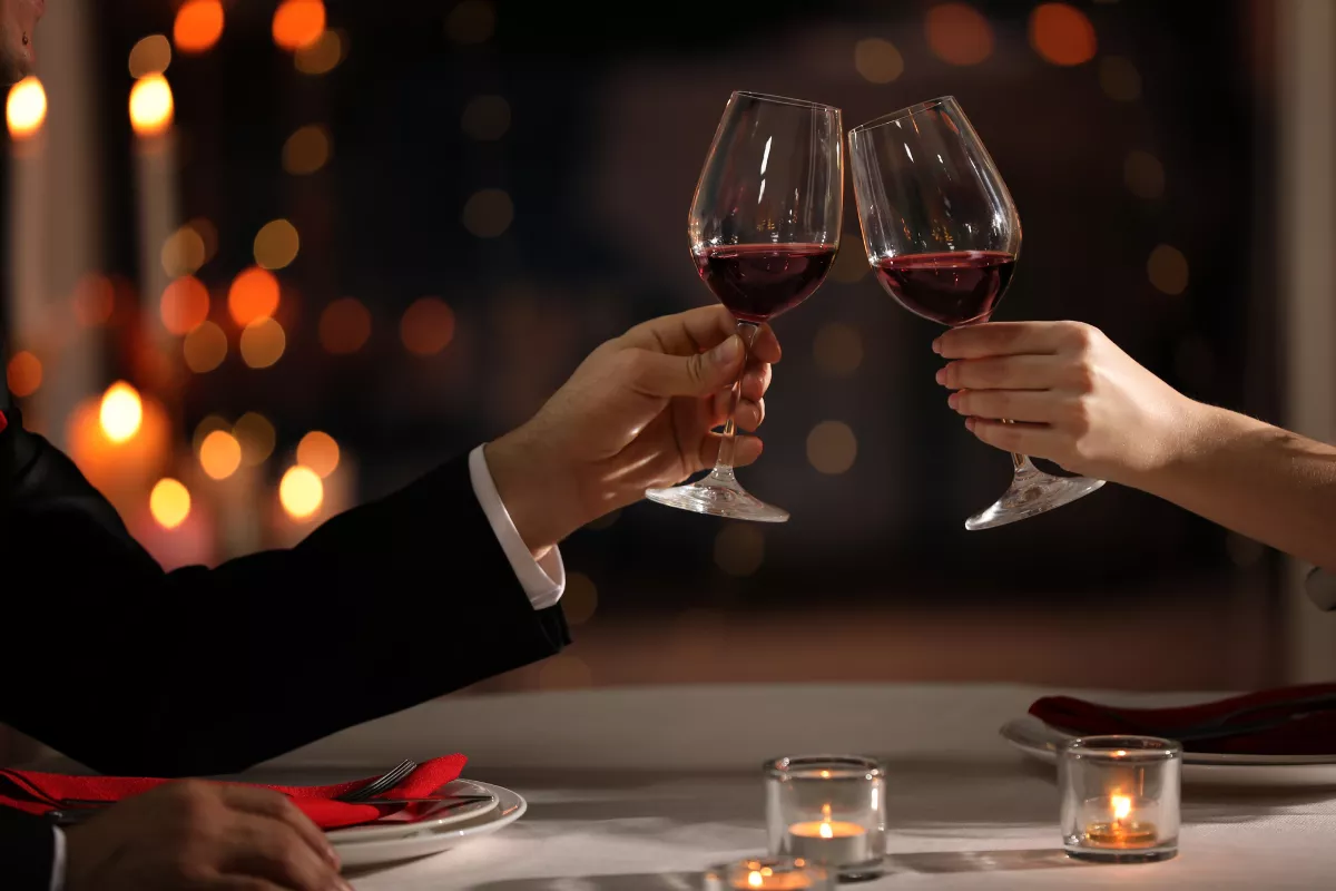 Casal brindando com taças de vinho tinto em mesa de jantar decorada com velas, representando um ambiente acolhedor e sofisticado para promoção especial de restaurante no Dia dos Namorados