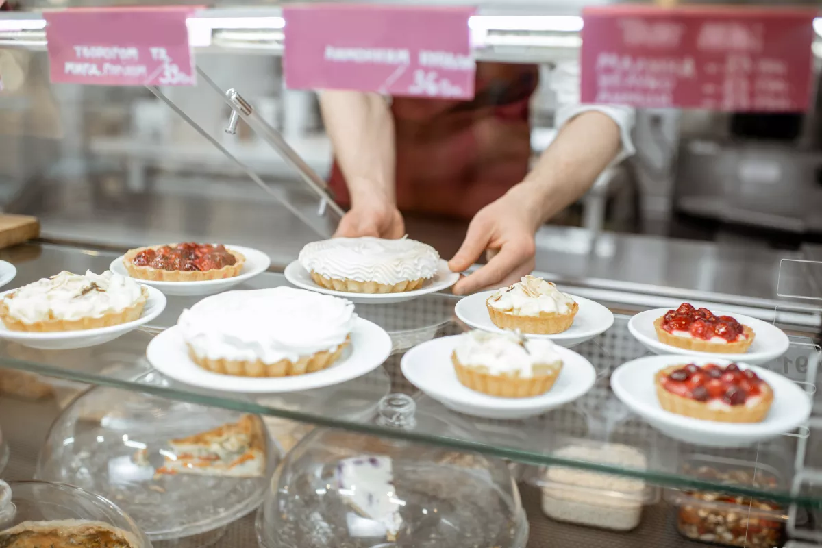 Doces artesanais dispostos em vitrine de loja física, com atendente servindo torta em prato branco, representando o atendimento presencial em confeitarias.