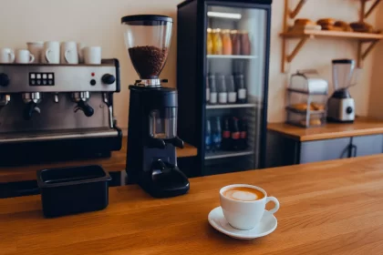 Máquina de café espresso profissional, moedor de grãos e geladeira expositora em bancada de cafeteria, com xícara de cappuccino em destaque.
