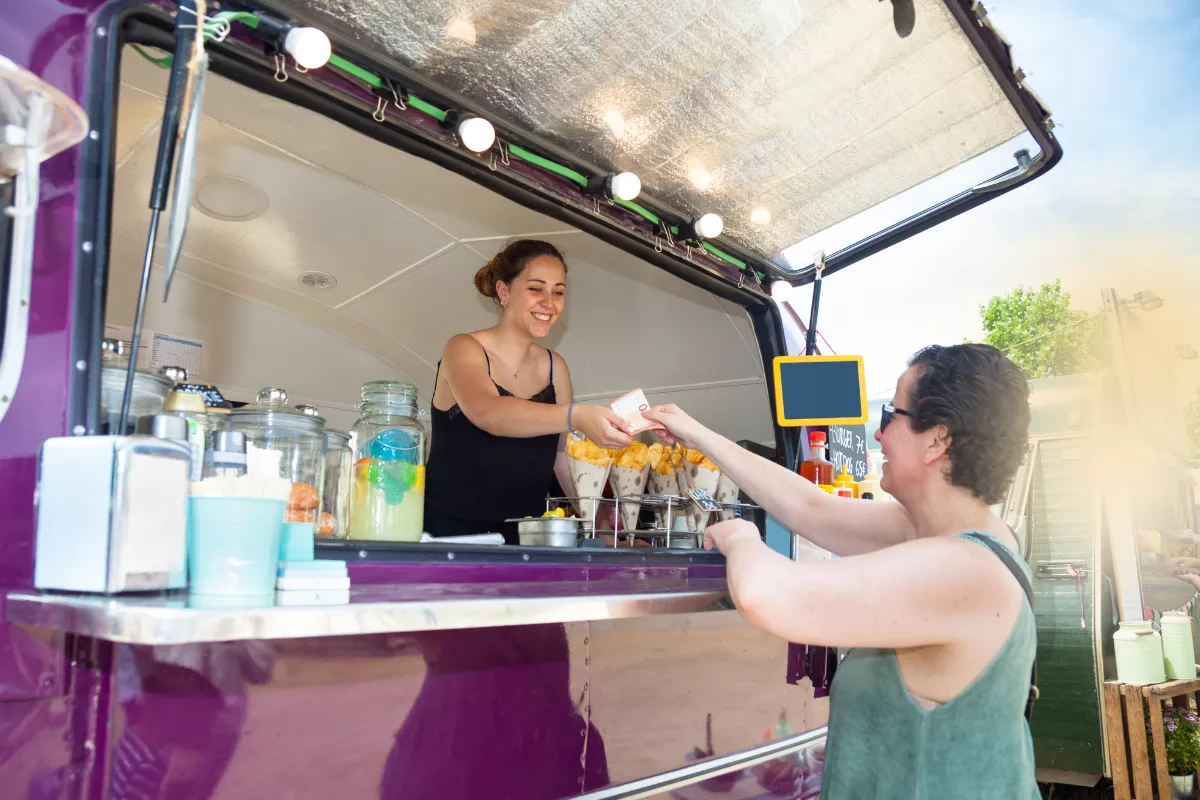 Atendente entrega pedido com sorriso a cliente em food truck roxo, com balcão repleto de bebidas e cones de batata, em ambiente ao ar livre.