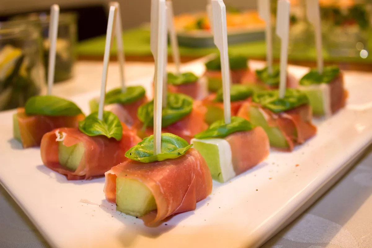 Finger food com cubos de melão e presunto cru, finalizados com folhas de manjericão e palitos, servidos em bandeja branca para eventos.