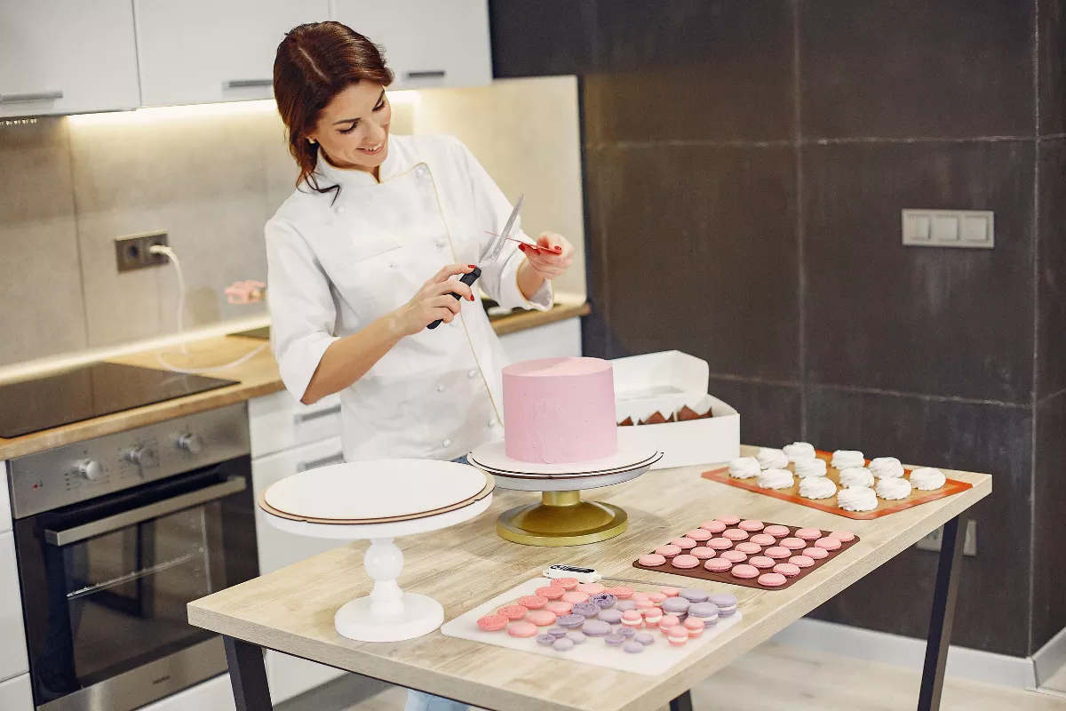 Confeiteira decorando um bolo rosa em cozinha caseira, rodeada por macarons e suspiros, ilustrando produção artesanal para confeitaria delivery.

