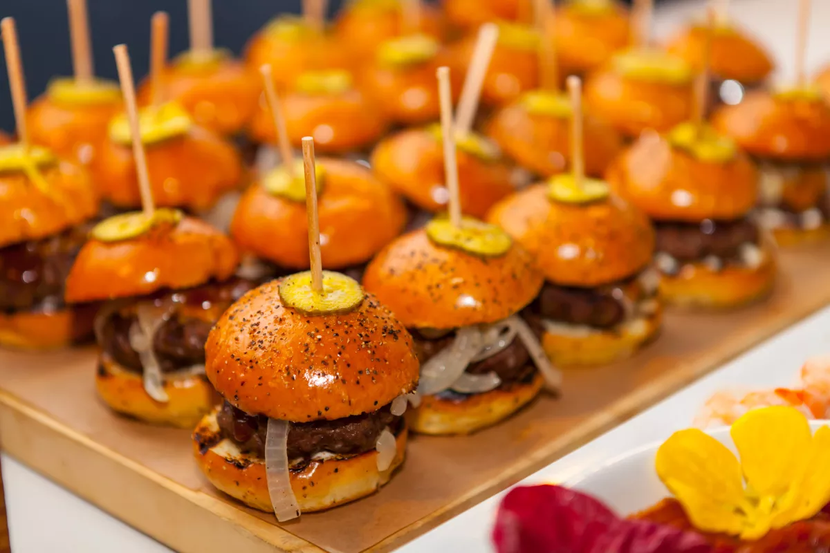 Mini hambúrgueres artesanais dispostos sobre uma bandeja, com pão dourado, carne, cebola e fatia de picles no topo, presos por palitos. Ideal para porções e eventos.