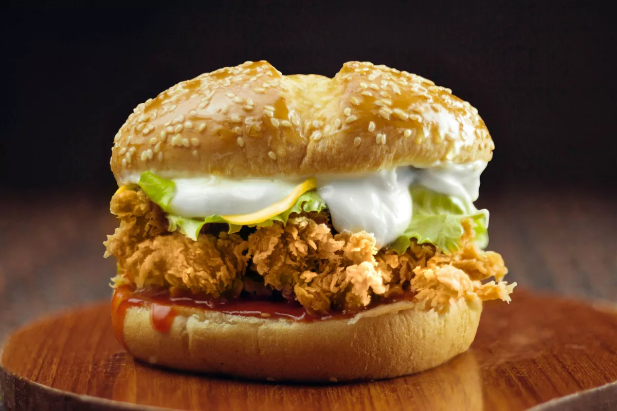 Hambúrguer de frango empanado e crocante, servido em pão de gergelim, com alface e maionese, sobre superfície de madeira. Alternativa leve e saborosa.