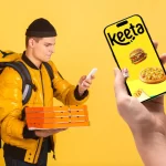 Entregador de delivery vestindo jaqueta amarela e mochila térmica segura caixas de pizza enquanto consulta o celular; em primeiro plano, uma mão segura um smartphone com a tela exibindo o app Keeta, ilustrado com imagens de hambúrguer, pizza e sorvete, sobre fundo amarelo vibrante.