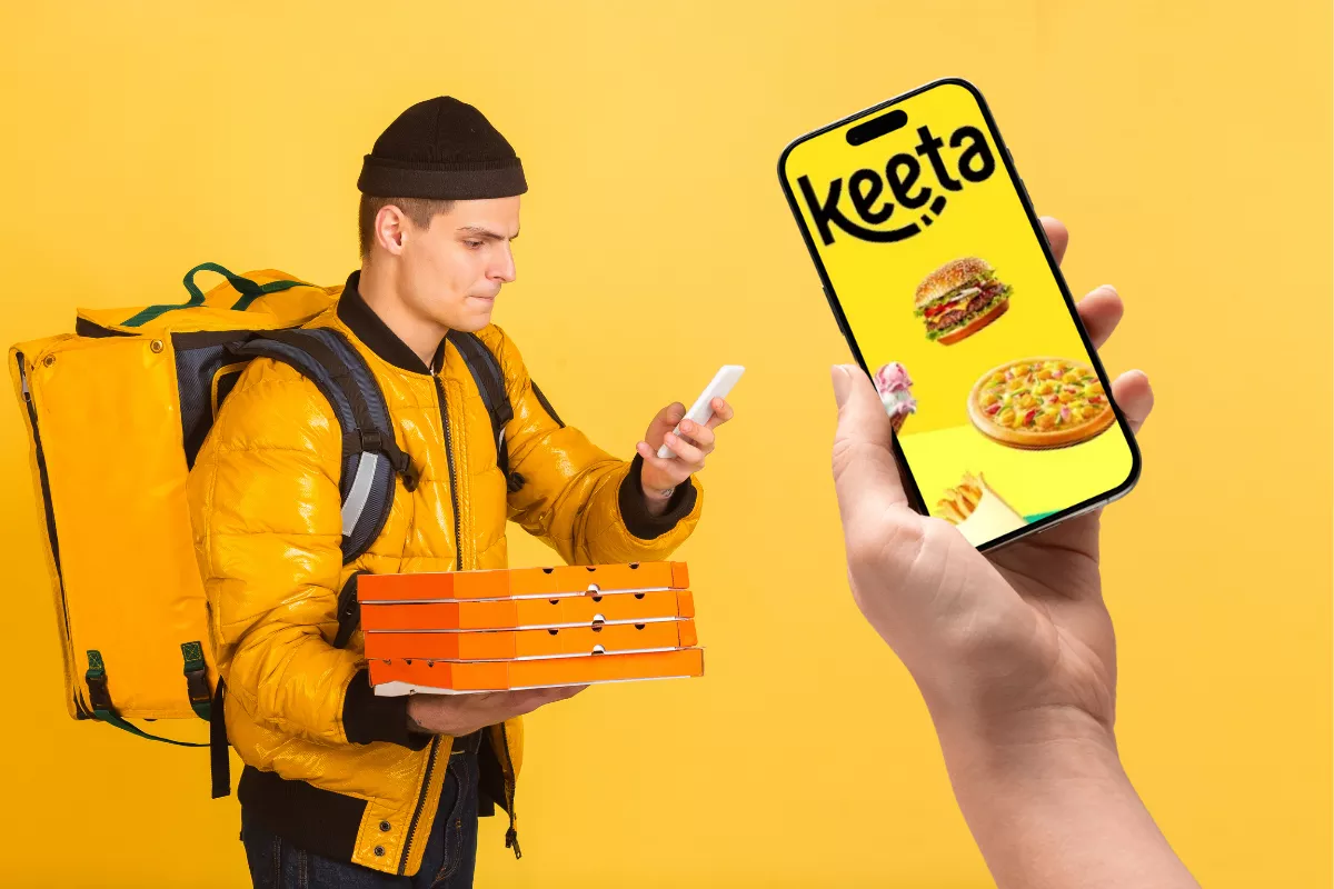 Entregador de delivery vestindo jaqueta amarela e mochila térmica segura caixas de pizza enquanto consulta o celular; em primeiro plano, uma mão segura um smartphone com a tela exibindo o app Keeta, ilustrado com imagens de hambúrguer, pizza e sorvete, sobre fundo amarelo vibrante.