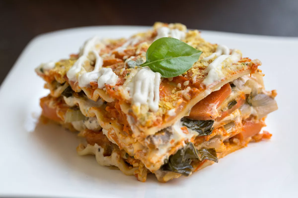 Porção de lasanha vegetariana com legumes, molho branco e folhas frescas, servida em prato branco, oferecendo uma opção inclusiva e saborosa para menus comemorativos.