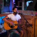 Cantor tocando violão e cantando ao vivo em barzinho aconchegante, com público próximo e ambiente informal — perfeito para uma playlist de música para barzinho.